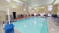 Located at 12710 N Mill Rd in Spokane, Washington, this single-story indoor aquatic facility is purpose-built for aquatic activities and therapy. The centerpiece of the space is a generously sized indoor swimming pool with clear, inviting water, bordered by wide concrete pool decks that provide ample space for movement and equipment. The facility features soaring vaulted ceilings with clerestory windows positioned high along the walls, allowing abundant natural light to flood the interior while maintaining a bright, airy atmosphere throughout. Large picture windows along the perimeter walls offer views of the surrounding wooded landscape, connecting the interior to the natural environment outside. The walls are finished with a warm beige tone, accented by a dark gray tile wainscoting that runs the length of the room, lending a clean and professional aesthetic. Safety features are prominently present, including a mounted life ring and accessible pool entry equipment such as a mechanical lift. A dedicated seating and waiting area with rattan-style chairs and small tables is integrated into the space near the entrance, ideal for observers or those awaiting use of the pool. Changing room access doors, a bench, and a water fountain station are also conveniently located within the facility. The overall design prioritizes functionality, accessibility, and comfort in a well-lit, spacious environment.