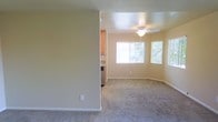 This single-floor space features an open-concept layout that connects the living area to the kitchen, creating a functional and cohesive flow. The living area is finished with wall-to-wall carpeting in a neutral gray tone, complementing the warm beige walls that run throughout. A ceiling fan with a light fixture is centrally positioned overhead, providing both ventilation and ambient lighting.

Two windows along the far wall allow natural light to fill the room, offering views of surrounding greenery and contributing to a bright, welcoming atmosphere. White baseboards provide a clean contrast against the carpeted floor and painted walls.

A partial wall separates the living area from the kitchen, which is visible in the background. The kitchen features warm wood-toned cabinetry and what appears to be stainless steel appliances, adding a practical and polished element to the space. The transition between rooms is straightforward, supporting ease of movement throughout the home.

Overall, the space presents a clean, move-in-ready environment with a simple, functional design characterized by neutral finishes, natural light, and an efficient single-floor layout.