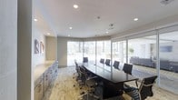 This single-floor professional meeting space is designed to accommodate collaborative work and formal gatherings with equal sophistication. At its center sits a large, dark-toned boardroom table surrounded by ergonomic high-back chairs, providing ample seating for sizable meetings or presentations. The room's most striking architectural feature is its expansive curved wall of floor-to-ceiling windows, which floods the interior with natural light and frames views of the surrounding outdoor area, including mature palm trees and an adjacent courtyard. The neutral color palette — soft whites, warm beiges, and subtle grey tones — creates a calm and focused atmosphere. Recessed lighting in the smooth white ceiling supplements the natural light, ensuring consistent illumination throughout. Built-in cabinetry along one wall offers practical storage solutions while maintaining the room's clean, uncluttered aesthetic. Patterned low-pile carpeting adds texture to the floor without distracting from the room's professional character. A mounted display on the wall further supports the space's function as a versatile, technology-ready meeting environment. Overall, the space strikes a balance between functionality and refined design.