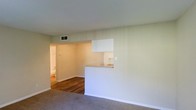 This single-floor space features an open, functional layout designed for comfortable everyday living. The main living area is finished with neutral-toned carpet, contributing to a warm and understated ambiance. Soft, light green walls run throughout, creating a cohesive and calming aesthetic across the space.

A defining architectural feature is the pass-through kitchen counter, topped with a light stone or laminate surface, which connects the kitchen to the living and dining area. White cabinetry is visible above the pass-through, offering practical storage while maintaining a clean, minimalist look. This open connection between the kitchen and living space promotes an airy, social atmosphere.

Beyond the main living area, a hallway leads to additional rooms, with hardwood-style flooring transitioning from the carpeted living space, adding visual contrast. Ceiling-mounted HVAC vents are integrated discreetly into the layout, ensuring year-round climate comfort. White baseboards frame the rooms cleanly, complementing the neutral wall tones. Overall, the space presents a straightforward, well-maintained interior with a practical, livable design.