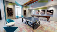 This dynamic, single-floor recreational space is thoughtfully designed to inspire energy and playfulness at every turn. The room's most eye-catching feature is its ceiling, adorned with bold, multicolored beams in teal, red, yellow, and green that add a striking artistic dimension overhead. Underfoot, a vibrant striped carpet in a rich palette of reds, blues, and neutrals anchors the space with equal visual flair.

At the center of the room stands a full-size Stiga ping-pong table, ready for competitive play, while a shuffleboard table along the back wall offers an additional gaming option. Colorful framed artwork and motorsport-themed prints decorate the walls, contributing to the room's lively and eclectic character.

Large windows along one side of the space invite an abundance of natural light, brightening the interior and providing views to the outside. Teal accent chairs and small side tables are arranged near the windows, creating a casual lounge area ideal for relaxing between games. Recessed and track lighting supplements the natural light, ensuring the space remains well-illuminated throughout.

Overall, this recreation room strikes a balance between energetic design and functional layout, making it a welcoming destination for social interaction, friendly competition, and leisure.
