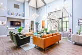 Located at 9 Lincoln Place in North Brunswick, NJ, this single-story community clubhouse offers an impressive and welcoming gathering space defined by its striking architectural design. The centerpiece of the space is a dramatic vaulted ceiling with exposed dark wood beams that rise to considerable heights, creating an airy, cathedral-like atmosphere that immediately commands attention upon entry.

The main lounge area is generously proportioned and bathed in natural light streaming through large arched glass doors and expansive windows that frame views of lush greenery outside. The interior is finished with wide-plank wood-look flooring in warm, neutral tones that complement the soft grey walls throughout. Contemporary Sputnik-style chandeliers add a sophisticated touch overhead.

Seating areas are thoughtfully arranged with modern furniture, including velvet accent chairs in olive green and mustard gold tones, accented by colorful throw pillows. A built-in refreshment station with white cabinetry and countertops is conveniently integrated into one wall, alongside a mounted flat-screen television. Curated artwork, potted plants, and decorative vases placed on elevated ledges contribute to a polished, inviting ambiance. Additional rooms accessible from the main area provide flexible spaces for varied activities, making this clubhouse a versatile and vibrant community hub.