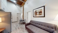 Located at 719 N Rowe St in Ludington, Michigan, this single-floor space showcases a compelling mix of industrial character and contemporary design. The living area immediately draws attention with its dramatically tall ceilings, exposed wooden timber beams, and visible ceiling structure, lending a loft-like atmosphere to the space. Light wood-tone flooring flows throughout, contributing to an open, airy feel. A striking exposed brick wall frames the entryway, where a dark-painted door with transom window allows natural light to filter in and adds a bold architectural focal point.

The living area comfortably accommodates a seating arrangement alongside a small dining table, making efficient use of the thoughtfully designed layout. The kitchen is outfitted with white shaker-style cabinetry, granite countertops, a classic subway tile backsplash, and stainless steel appliances including a range, dishwasher, and over-the-range microwave.

The bedroom is a calm, well-proportioned retreat with light walls, wood-tone flooring, and a window providing natural light. The bathroom mirrors the kitchen's refined aesthetic, featuring a white vanity with granite countertop, a large mirror, and wood-tone tile flooring. Throughout, the space balances historic architectural details with clean, modern finishes.