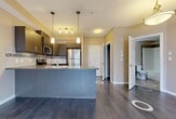 Located in Edmonton, Alberta, this well-designed one-bedroom, two-bathroom unit offers a practical and modern layout across a single floor. The open-concept living and kitchen area is anchored by rich dark hardwood-style flooring and warm neutral-toned walls, creating a cohesive and inviting atmosphere. The kitchen is a standout feature, equipped with a full suite of stainless steel appliances including a refrigerator, electric range, built-in microwave, and dishwasher. Dark wood cabinetry is complemented by a decorative mosaic tile backsplash, granite-style countertops, and a peninsula island with pendant lighting overhead — ideal for casual dining or meal prep. The living area benefits from a sliding glass door that allows natural light to flow in. The bedroom is comfortably sized, finished with plush carpeting, a window with exterior views, and a closet with built-in shelving. Two bathrooms serve the unit — one featuring a full tub-shower combination, dark wood vanity, granite-style countertop, and stone-look tile flooring. An additional tiled space provides utility storage. The overall design balances functionality with contemporary style, making this a well-appointed urban residence.