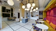 The lobby at 800 Tower makes an immediate impression with its bold, carefully curated design aesthetic. Large-format marble tile flooring stretches across the open space, reflecting the abundant light from a dramatic cluster of oversized globe pendant lights suspended from a geometric black metal framework overhead. The ceiling height lends the space an airy, grand atmosphere that sets a welcoming tone from the moment of arrival.

A central reception desk anchors the space, finished in a sleek combination of marble and soft grey tones, positioned beneath the striking light installation. Gold-patterned textured columns add warmth and visual interest, complementing the overall color palette of white, gold, and rich jewel tones found throughout the furnishings.

Seating areas are thoughtfully arranged to encourage both social interaction and relaxed waiting. Floral-upholstered accent chairs in deep purples and blues are paired with round white-topped bistro tables, while a classically styled sofa with colorful patterned fabric adds an eclectic, artistic touch. A flat-screen display and additional lounge seating are visible toward the rear, rounding out the functional amenities of this inviting space. The overall ambiance blends contemporary elegance with artistic flair, creating a lobby that is both distinctive and welcoming.