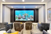 Located at 12389 Merit Dr in Dallas, TX, this stylish media lounge is a compact yet thoughtfully designed space that delivers an upscale viewing and relaxation experience. The focal point of the room is a large, multi-panel video wall set within a dark, textured surround featuring a geometric hexagonal tile pattern, creating a striking cinematic backdrop. The display is currently showcasing a vibrant underwater aquarium scene, adding a dynamic, immersive quality to the ambiance.

Seating consists of a collection of plush, gray upholstered armchairs accented with bold yellow throw pillows, arranged to face the feature wall. Low, cylindrical white accent tables are positioned throughout the seating area for convenience. The floor is covered in a neutral, patterned area rug that adds texture and warmth.

The ceiling design is a standout architectural element, featuring layered coffers with integrated LED strip lighting that casts a soft, warm glow throughout the space. Recessed downlights provide additional illumination, contributing to the room's polished, contemporary feel. Along one wall, tall arched niches display framed botanical artwork, further enhancing the refined aesthetic. The entryway opens to a broader corridor with glass double doors, allowing natural light to filter in and connect the lounge to the surrounding facility.