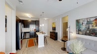 This single-floor residence offers a thoughtfully designed open-concept layout that seamlessly connects the kitchen, dining, and living areas into one cohesive, light-filled space. The kitchen is a standout feature, boasting rich dark cabinetry that contrasts beautifully with a classic white subway tile backsplash and light countertops. A central kitchen island with bar seating provides both a functional prep space and a casual dining option, illuminated by elegant pendant lights overhead. Stainless steel appliances, including a refrigerator and dishwasher, complete the well-appointed kitchen. Light wood-tone flooring flows throughout the main living area, contributing to a warm and welcoming ambiance. The living space is open and airy, with a ceiling fan and contemporary industrial-style floor lamp adding character. A hallway leads to additional rooms beyond, offering privacy and separation from the main living areas. Neutral white walls and high ceilings throughout the space enhance the sense of openness, creating a bright and inviting atmosphere that is both functional and stylish.