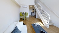 This two-story loft offers a well-designed open-concept layout that seamlessly connects the living, dining, and kitchen areas on the main floor. Warm hardwood flooring runs throughout the space, contributing to a clean and cohesive aesthetic. The living area features a white sofa and a sleek coffee table, creating a comfortable and minimalist seating arrangement. Adjacent to the living space, a compact round dining table paired with vibrant lime-green chairs adds a lively pop of color to the otherwise neutral palette.

The kitchen is efficiently laid out along the back wall, equipped with stainless steel appliances including a refrigerator and microwave, complemented by dark countertops and white cabinetry. A wall-mounted split air conditioning unit ensures year-round climate control. Natural light brightens the main floor, enhancing the open feel of the space.

A white metal-railed staircase rises prominently along one wall, leading to the upper level, which overlooks the main living area through an open balustrade. The double-height ceiling above creates a strong sense of volume and airiness, making the overall space feel expansive and modern.