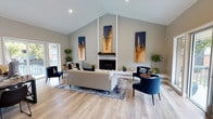 This thoughtfully designed single-floor space combines elegant aesthetics with practical functionality. The centerpiece of the main living area is a striking floor-to-ceiling white brick fireplace, flanked by three large navy and gold abstract art panels that draw the eye upward toward the dramatic vaulted ceiling. Recessed lighting and large sliding glass doors flood the space with natural light, offering views of and direct access to an outdoor balcony area.

Light hardwood flooring runs throughout, complementing the neutral gray-toned walls and lending warmth to the interior. The living area is furnished with a generous sectional sofa, an abstract area rug in navy and cream, and a pair of deep blue velvet accent chairs, creating a sophisticated and cohesive color palette.

Adjacent to the main living area, a dedicated workspace features a dark-toned desk and office seating, providing a functional professional zone within the open-plan layout.

A separate kitchenette area offers navy cabinetry with light marble-style countertops, a built-in beverage refrigerator, a stainless steel microwave, and a sink. A nature-themed birch forest wall mural serves as a distinctive focal point, adding a creative and refreshing element to the space. Overall, the design balances contemporary style with inviting comfort.