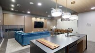 Located at 590 Park St in St. Paul, Minnesota, this single-floor amenity space offers a seamlessly integrated open-plan layout that combines a full kitchen, lounge, and dining area within a well-appointed, contemporary design. The kitchen is a standout feature, anchored by a large dark island with bar seating and an integrated sink beneath a cluster of globe pendant lights with brass hardware. A striking blue herringbone tile backsplash frames a professional-grade range and hood, complemented by light wood cabinetry, open shelving, and a full-size refrigerator. A dramatic abstract mural in muted purples, grays, and blues serves as a bold focal point, bridging the kitchen and lounge zones. The lounge area is furnished with plush blue velvet sofas and swivel chairs arranged around a built-in wood media wall housing a large flat-screen television and a sleek linear fireplace. A wood-paneled ceiling detail adds warmth over the kitchen, while recessed lighting and in-ceiling speakers provide a refined ambiance throughout. A dining area with a long table and upholstered chairs rounds out the space, making it well-suited for both casual gatherings and more formal entertaining. Wide-plank hardwood-style flooring unifies the entire layout with understated elegance.