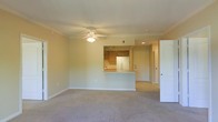 This single-floor space features an open-concept layout that seamlessly connects the living area to the kitchen. The living room is finished with wall-to-wall neutral carpet and painted in soft, warm tones, creating a bright and welcoming atmosphere. Crown molding runs along the ceiling perimeter, adding a refined architectural detail, while a ceiling fan with a light fixture provides both comfort and illumination.

The kitchen is accessible via a half-wall pass-through counter, which also serves as a breakfast bar. Warm wood-toned cabinetry lines the kitchen walls, complemented by a white refrigerator and track lighting overhead, offering functional and efficient use of the space. The pass-through design enhances the open feel while maintaining a defined cooking area.

A doorway visible from the main living area leads to additional rooms or a hallway, suggesting further private spaces beyond the main living zone. A white panel door on the right side of the living area provides access to what appears to be a closet or utility space. Overall, the unit presents a clean, move-in-ready environment with consistent neutral finishes throughout.