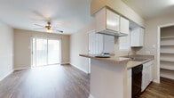 This single-story, 1-bedroom, 1-bathroom residence features a practical and cohesive layout with wood-style plank flooring carried throughout. The living area is bright and open, benefiting from a sliding glass door with vertical blinds that leads to a private balcony, allowing natural light to fill the space. A ceiling fan is centrally positioned overhead, providing both airflow and ambient lighting.

The galley-style kitchen is efficiently designed with white cabinetry lining both sides, black appliances including a range and dishwasher, a single-basin sink, and laminate countertops. A peninsula with a raised bar counter opens toward the dining and living areas, creating a connected feel. A built-in open shelving unit at the end of the kitchen provides additional storage.

The dining area is defined by a pendant light fixture and a decorative chair rail that runs along the neutral-toned walls. The bedroom is a comfortable, quiet space with a ceiling fan and a window that allows natural light. The bathroom includes a vanity with storage and a full tub-shower combination. The unit offers a functional, straightforward layout well-suited for everyday living.