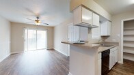 This single-story, 1-bedroom, 1-bathroom residence features a practical and cohesive layout with wood-style plank flooring carried throughout. The living area is bright and open, benefiting from a sliding glass door with vertical blinds that leads to a private balcony, allowing natural light to fill the space. A ceiling fan is centrally positioned overhead, providing both airflow and ambient lighting.

The galley-style kitchen is efficiently designed with white cabinetry lining both sides, black appliances including a range and dishwasher, a single-basin sink, and laminate countertops. A peninsula with a raised bar counter opens toward the dining and living areas, creating a connected feel. A built-in open shelving unit at the end of the kitchen provides additional storage.

The dining area is defined by a pendant light fixture and a decorative chair rail that runs along the neutral-toned walls. The bedroom is a comfortable, quiet space with a ceiling fan and a window that allows natural light. The bathroom includes a vanity with storage and a full tub-shower combination. The unit offers a functional, straightforward layout well-suited for everyday living.