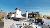 The Kam Building Rooftop at 918 E Thompson Blvd in Ventura, California, is a thoughtfully designed amenity space that seamlessly blends indoor comfort with expansive outdoor living. The rooftop terrace is finished with large-format porcelain tile and anchored by a striking wood-beam pergola that frames sweeping views of the Pacific Ocean, palm tree-lined streets, and the surrounding Ventura hillside. Beneath the pergola, a generous outdoor lounge area features contemporary sectional seating and dining tables, ideal for gathering and entertaining. A concrete bar counter with metal barstools provides an additional casual seating option, while a central fire pit surrounded by modern lounge chairs creates an inviting focal point for evening use. A dedicated recreation zone offers a foosball table and ping-pong table for added entertainment. The interior portion of the rooftop houses a bright, modern lounge with large windows and glass doors that flood the space with natural light and maintain visual connectivity to the outdoor deck. This indoor area includes a living room setup with a wall-mounted television, leather seating, and a compact dining nook. A kitchenette equipped with dark cabinetry, white countertops, subway tile backsplash, and stainless steel appliances rounds out the interior amenities, making this rooftop a fully functional and stylish communal destination.
