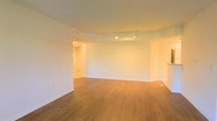 This single-floor space presents a clean, modern aesthetic defined by warm hardwood flooring, crisp white walls, and a smooth, well-maintained ceiling throughout. The main living area is generously proportioned, offering an open-plan layout that lends itself to flexible use and easy customization. A track lighting system is mounted along the ceiling, providing directed, adjustable illumination that enhances the overall brightness of the interior. The curved architectural detail along the ceiling adds a subtle yet distinctive design element, giving the space a sense of refined character. White baseboards provide a polished finish along the perimeter of the room. A connecting hallway is visible toward the rear of the main area, leading to additional rooms and the entry, suggesting a functional and well-organized floor plan. The neutral color palette throughout — from the flooring to the walls and ceiling — creates a cohesive, airy ambiance that feels welcoming and easy to personalize. The overall atmosphere is bright, clean, and well-appointed, making this a practical and visually appealing single-floor layout suitable for a variety of uses.