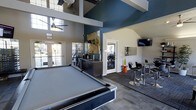 This single-floor amenity lounge is designed to serve as a dynamic social and recreational hub. The space immediately impresses with its soaring vaulted ceilings accented by bold teal-blue tones and crisp white structural beams, creating an open and energetic atmosphere. A full-size billiards table anchors the main area, making it a natural focal point for recreation and socializing. Large windows and glass-paneled doors along the perimeter flood the interior with abundant natural light while offering views to the exterior. A ceiling fan ensures comfortable airflow throughout the open layout. Adjacent to the billiards area, a stylish lounge zone features modern bar-height seating arranged around a high-top table, accompanied by a wall-mounted flat-screen TV — ideal for watching sports or casual gatherings. A bold abstract painting adds an artistic touch to the wall, complementing the contemporary color palette. A dark, built-in credenza provides functional storage while reinforcing the sleek, modern aesthetic. Through a doorway, a dedicated fitness area is visible, equipped with exercise machines, offering residents convenient access to workout facilities. The combination of recreational, social, and fitness amenities within this thoughtfully designed space makes it a versatile and inviting communal destination.