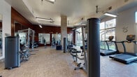 This fitness center offers a spacious, thoughtfully designed workout environment spread across a single floor. The space is divided into distinct functional zones, accommodating a wide range of exercise disciplines. The strength training area features cable machines, multi-use weight stations, a Concept2 rowing machine, and free weights, complemented by floor-to-ceiling mirrors that visually expand the space. Warm wood-toned accent walls and exposed brick columns add architectural character, balancing the industrial aesthetic with welcoming warmth.

The cardio zone is well-appointed with TRUE brand treadmills, elliptical trainers, stationary bikes, and stair climbers, all positioned to take advantage of the abundant natural light streaming through large floor-to-ceiling windows. These windows also provide direct views of and access to an adjacent outdoor turf area, seamlessly connecting indoor and outdoor training options.

A dedicated functional training corner includes plyo boxes, medicine balls, stability balls, and a wall-mounted rack, catering to dynamic and cross-training workouts. Ceiling fans and pendant lighting maintain comfort throughout. The durable, easy-to-maintain flooring and high ceilings contribute to an open, airy ambiance suited for high-energy workouts.