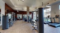 This fitness center offers a spacious, thoughtfully designed workout environment spread across a single floor. The space is divided into distinct functional zones, accommodating a wide range of exercise disciplines. The strength training area features cable machines, multi-use weight stations, a Concept2 rowing machine, and free weights, complemented by floor-to-ceiling mirrors that visually expand the space. Warm wood-toned accent walls and exposed brick columns add architectural character, balancing the industrial aesthetic with welcoming warmth.

The cardio zone is well-appointed with TRUE brand treadmills, elliptical trainers, stationary bikes, and stair climbers, all positioned to take advantage of the abundant natural light streaming through large floor-to-ceiling windows. These windows also provide direct views of and access to an adjacent outdoor turf area, seamlessly connecting indoor and outdoor training options.

A dedicated functional training corner includes plyo boxes, medicine balls, stability balls, and a wall-mounted rack, catering to dynamic and cross-training workouts. Ceiling fans and pendant lighting maintain comfort throughout. The durable, easy-to-maintain flooring and high ceilings contribute to an open, airy ambiance suited for high-energy workouts.