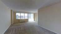 This single-floor space features an open-plan living area designed to maximize both light and usability. The room is finished with wall-to-wall neutral-toned carpeting that extends throughout, providing a soft, understated foundation. Walls are painted in a warm cream tone, contributing to a bright and airy atmosphere.

A prominent feature of the living area is the large picture window paired with a floor-to-ceiling vertical blind installation, allowing for flexible light control while framing an elevated city skyline view. The windows also reveal a baseboard heating unit positioned beneath, offering an efficient heating solution integrated into the design.

The ceiling is smooth and flat, finished in a light tone that reflects natural light effectively. The layout is open and expansive, offering considerable flexibility for furniture arrangement. Clean white baseboards line the perimeter of the room, adding a crisp contrast to the carpeting. The overall ambiance is neutral, practical, and well-lit, making it a versatile space well-suited for a variety of uses.