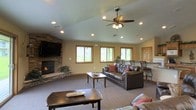 This single-story space centers around a spacious, open living area defined by a vaulted ceiling that lends an airy, expansive feel to the room. Recessed lighting is embedded throughout the ceiling and is complemented by a ceiling fan with an integrated light fixture, ensuring the space is well-lit at any time of day. A corner stone fireplace serves as a prominent focal point, featuring stacked natural stonework that extends to the wall and provides a rustic, warm aesthetic. A wall-mounted flat-screen television is positioned above the fireplace, adding a functional entertainment element to the space. The living area is carpeted and comfortably furnished with dark leather seating accented by warm-toned throw pillows, arranged around a pair of wooden coffee tables. Multiple windows with warm wood trim line the walls, flooding the interior with natural light and offering views of the surrounding greenery outdoors. A bar-height seating area with tall chairs is visible toward the back of the room, adding a casual dining or socializing nook. Cabinetry with natural wood finishes can also be seen in the background, contributing to the cohesive, warm design throughout the space.