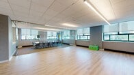 This contemporary single-floor office space is thoughtfully designed to accommodate both collaborative and focused work. The open-plan area features light hardwood flooring that creates a clean, bright atmosphere, complemented by a drop-tile ceiling with integrated linear LED lighting that provides consistent illumination throughout. Textured blue-gray accent walls add depth and a modern aesthetic to the interior.

A standout feature of the space is the glass-walled conference room, enclosed by floor-to-ceiling frameless glass panels that maintain visual openness while offering a defined meeting environment. The conference room is furnished with a large rectangular table surrounded by ergonomic rolling chairs, and is carpeted in a warm neutral tone. Multiple windows with roller shades line the conference room walls, offering views of the surrounding cityscape and flooding the area with natural light.

The open area adjacent to the conference room is flexible and generously proportioned, suitable for a variety of uses. Large windows along the perimeter bring in abundant natural light and frame outdoor views of the urban skyline. Additional utility panels and control interfaces are mounted on the walls, and a private door provides access to a separate room. The overall design balances functionality and modern aesthetics, making it a versatile and professional environment.
