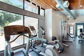 Located at 1901 Briarcliff Rd NE in Atlanta, Georgia, this fitness center offers a well-appointed exercise space designed with both function and aesthetics in mind. The facility is organized into two connected zones that flow seamlessly into one another, providing dedicated areas for cardio and strength training. The cardio section is highlighted by dramatic floor-to-ceiling glass walls that flood the space with natural light and offer direct views of the outdoor pool and surrounding lush greenery, creating an energizing workout environment. A warm natural wood-planked ceiling with exposed industrial-style HVAC ductwork adds architectural character to this area, complemented by track lighting overhead. The strength training zone features durable rubber flooring, a multi-station cable machine, an adjustable weight bench, and a full rack of dumbbells for versatile free-weight workouts. Cardio equipment includes treadmills, elliptical trainers, and a stationary bike. Softer blue-gray painted walls with recessed lighting define the strength area, while a framed art piece adds a refined touch to the otherwise utilitarian setting. Windows along the perimeter of the strength zone overlook wooded surroundings, maintaining a connection to nature throughout the space. The fitness center combines contemporary design with practical amenities to deliver a comprehensive workout facility.