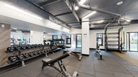 This amenity space comprises a generously sized fitness center and a dynamic outdoor courtyard, offering a well-rounded environment for both exercise and leisure. The fitness center is divided into distinct workout zones. The main training area features rubber flooring, exposed ductwork overhead, and industrial-style ceiling lighting that gives the space a modern, utilitarian aesthetic. It is equipped with a comprehensive free weights section, including a full dumbbell rack, kettlebells, adjustable benches, and a functional training rig with resistance bands. A separate cardio room houses treadmills, elliptical machines, and stationary cycling bikes, all positioned near windows that let in natural light and provide views of the outdoor area. A sauna or ancillary room is also accessible from the cardio zone. The outdoor courtyard is a standout feature, offering a lively and inviting atmosphere framed by exposed brick walls and modern architectural cladding. Concrete raised planters are filled with trees and shrubs, while a bold rainbow-striped rubber surface anchors a seating area with colorful Adirondack-style chairs. A nostalgic vintage "Car Wash" marquee sign adds a playful, artistic focal point. Multiple gas BBQ grills and brightly colored dining furniture make this an ideal space for outdoor gatherings.