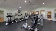 This fitness center is thoughtfully designed to support a full range of workout routines in a comfortable, well-appointed environment. The space features vaulted ceilings with recessed lighting and a ceiling fan that contribute to an open, airy atmosphere. Soft green walls and crown molding lend the room a polished finish rarely found in typical gym settings.

A standout architectural detail is the natural stone accent wall surrounding a fireplace, adding warmth and character to the space. Large French doors and multi-pane windows along the perimeter flood the interior with natural light and offer views of the outdoors, creating an inviting workout environment.

The cardio zone is well-stocked with multiple treadmills, elliptical trainers, and stationary and recumbent bikes — all from True Fitness — ensuring ample options for aerobic exercise. A wall-mounted flat-screen television is positioned for easy viewing during cardio sessions. The strength training area features a cable crossover machine, selectorized weight machines, free weights, and stability balls to accommodate a variety of training styles.

Dark rubber flooring throughout provides durability and shock absorption. A large wall mirror enhances the sense of space and supports proper form during exercise. The fitness center is completed with safety features including clearly marked emergency exits, a fire extinguisher, and security camera coverage.