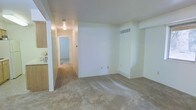 This single-floor space presents a straightforward, functional layout centered around a spacious main living area. The room is finished with wall-to-wall carpeting in a neutral beige tone, providing a soft underfoot surface throughout. White walls keep the interior bright and versatile, offering a blank canvas for various furnishing arrangements.

A visible HVAC vent and baseboard unit along the far wall indicate climate control capabilities within the living space. Electrical outlets are positioned at standard intervals along the walls, ensuring practical usability.

A hallway extends from the main living area, illuminated by a ceiling-mounted light fixture, and leads toward additional rooms visible in the distance, suggesting a private sleeping area or bathroom beyond. The hallway's carpeting continues seamlessly from the main space, maintaining a consistent aesthetic throughout.

To the left, a partial view of what appears to be a kitchen area is visible, featuring wooden cabinetry, hinting at a functional cooking space adjacent to the living room. The overall design is clean, minimal, and utilitarian, with a neutral color palette that lends itself well to personalization.