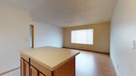 This single-floor unit is designed with an open and functional layout. The kitchen serves as a central hub, featuring warm oak cabinetry that extends to the upper walls for ample storage, a peninsula with a laminate countertop that doubles as a breakfast bar, and white appliances including a refrigerator, electric range, dishwasher, and sink. Tile flooring grounds the kitchen area, transitioning into light wood-style laminate flooring that continues throughout the living spaces.

The open-concept design connects the kitchen directly to the living area, which is generously sized and filled with natural light from a large window fitted with horizontal blinds. A baseboard heating unit runs beneath the window, and the textured ceiling adds a classic residential character to the space.

Additional rooms feature the same light laminate flooring, neutral wall tones, and textured ceilings, offering versatile spaces suitable for various uses. The bathroom includes an oak vanity with a single sink, laminate countertop, and ample cabinet storage below, complemented by a tile floor. A full tub-shower combination with white tile surround completes the bathroom. Overall, the unit presents a clean, neutral palette throughout.