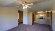 This single-floor residence offers a functional and practical layout designed for comfortable everyday living. The main living area is finished with wall-to-wall carpet in a neutral tone, creating a warm and inviting atmosphere. A ceiling fan with a built-in light fixture is centrally positioned overhead, providing both airflow and ambient lighting throughout the space.

The living area flows seamlessly into the kitchen, which is defined by warm wood-toned cabinetry and a light-colored countertop that doubles as a breakfast bar or pass-through surface. The kitchen is equipped with standard appliances, including a refrigerator and a range, and is illuminated by overhead fluorescent lighting. The light flooring in the kitchen contrasts with the carpeted living space, helping to visually distinguish each functional zone.

A hallway extends from the main living area, leading to additional rooms within the unit. A closet door is conveniently located near the transition between the living and kitchen areas, offering accessible storage. The overall layout is straightforward and efficient, with neutral wall tones and consistent finishes throughout that lend the space a clean, understated character suitable for a variety of interior styles.