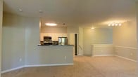 Located at 2600 Harvest Creek Pl in Cary, NC, this single-floor residence offers a thoughtfully designed open-concept layout that connects the kitchen and living areas seamlessly. The kitchen is well-appointed with stainless steel appliances, including a refrigerator and microwave, complemented by crisp white cabinetry and a dark tile backsplash that adds a modern contrast. A peninsula-style breakfast bar with a dark countertop extends into the living space, making it ideal for casual dining or entertaining. The living area features plush wall-to-wall carpeting in a neutral tone, contributing to a warm and comfortable ambiance. Soft, neutral-painted walls throughout enhance the brightness of the space, while flush-mounted ceiling lights provide consistent illumination. A staircase area is visible near the living space, and a doorway leading to additional areas hints at further functional spaces beyond. The overall design is clean, modern, and functional, making it a practical and inviting home in the heart of Cary, NC.