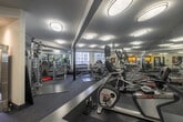 Located at 8665 Burton Way in Los Angeles, California, this fitness center offers a thoughtfully designed workout space across a single floor. The room is finished with patterned carpet flooring and features a clean, modern aesthetic with neutral-toned walls and a tiered ceiling with recessed sections accented by flush-mount circular light fixtures that provide consistent, even illumination throughout.

Floor-to-ceiling mirrors line multiple walls, visually expanding the space and enhancing the open, airy feel of the room. A standout decorative element is a freestanding stainless steel water feature near the entrance, adding a calming, upscale ambiance to the fitness environment.

The facility is well-appointed with a comprehensive range of equipment, including treadmills, elliptical trainers, rowing machines, cable machines, and a dumbbell rack, catering to both cardio and strength training needs. Large exercise mats are laid out in a dedicated stretching and floor exercise area. Structural columns with vertical poles are integrated throughout the space.

Near the entrance, a built-in cabinetry station with counter space, open shelving, and a mini-refrigerator provides convenient access to refreshments. A framed art piece and a mosaic tile accent wall add decorative character. Natural light filters in through windows visible in the background, complementing the overall well-lit and welcoming atmosphere.