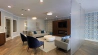 This single-floor space presents a thoughtfully designed lounge area that balances comfort and contemporary style. The room centers around a sleek, linear fireplace set into a rich, dark wood-paneled accent wall, above which a wall-mounted flat-screen television is installed. Adjacent to the accent wall, a bold patterned wallcovering in blue and white adds visual interest and texture, serving as a distinctive design element that anchors the space.

The seating arrangement is carefully curated, featuring a light gray sofa adorned with blue and white accent pillows, complemented by a matching gray accent chair and a pair of deep navy blue lounge chairs with dark wood legs. Organic-shaped white coffee tables sit atop a neutral area rug, adding a sculptural quality to the layout. The wood-look flooring throughout lends warmth to the otherwise cool color palette.

Recessed lighting and flush-mount ceiling fixtures provide even illumination throughout the space. A glimpse into an adjoining area suggests additional functional zones within the layout. The overall ambiance is polished and inviting, suitable for relaxation or social gathering.