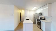 This single-floor residence offers a thoughtfully designed open-concept layout with a seamless flow between the living and kitchen areas. Light wood-style flooring runs throughout the main living space, contributing to a clean and contemporary aesthetic, while a modern chandelier fixture adds a subtle decorative touch overhead. A sliding glass door connects the living area to an outdoor space, allowing natural light to fill the room.

The kitchen is a standout feature, centered around a peninsula with a white quartz countertop and an integrated sink. Crisp, high-gloss white cabinetry extends to the ceiling for ample storage, complemented by a classic white subway tile backsplash. Stainless steel appliances include a gas range, over-the-range microwave, refrigerator, and dishwasher, providing a fully equipped cooking environment.

Two bedrooms are finished with plush carpeting and neutral walls. One bedroom features a sliding-door closet with built-in hanging rods, while the other benefits from a large window with vertical blinds, admitting natural light and outdoor views.

The bathroom is clean and functional, featuring a combined tub and shower with a curved curtain rod, a vanity with a light stone-topped countertop, and a framed mirror.