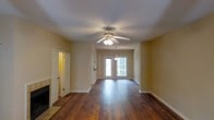 This single-floor residence offers a welcoming and functional layout designed for comfortable living. The main living area is a standout feature, showcasing rich, dark hardwood flooring that runs throughout and adds warmth and character to the space. A ceiling fan with a built-in light fixture is centrally positioned overhead, ensuring both comfort and illumination. A tiled fireplace anchors one side of the room, providing a classic focal point and a cozy atmosphere. Natural light pours in through a pair of glass-paneled front doors, brightening the interior and offering a pleasant connection to the outdoors. The neutral, warm-toned walls complement the flooring and contribute to an inviting ambiance throughout. A doorway on the opposite side of the living room leads to an adjacent space — likely a hallway or secondary room — where additional lighting is visible, hinting at a thoughtfully connected interior layout. White trim and baseboards provide clean contrast against the walls and floors, giving the space a polished, well-maintained appearance. Overall, this residence combines classic design elements with practical features, creating a comfortable and visually cohesive single-floor living environment.