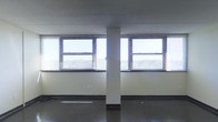 This single-floor space presents an open, functional layout well-suited for commercial or office use. The room features dark vinyl flooring that contrasts with the light-colored walls, creating a clean and utilitarian aesthetic. A central structural column divides the space visually without fully interrupting the open floor plan, adding an architectural element to the otherwise straightforward layout. Two large windows fitted with horizontal blinds flank the central column, allowing ample natural light to filter into the interior and providing views of the surrounding outdoor environment. The windows are framed with dark trim, offering a subtle contrast against the pale walls. The ceiling is flat and finished in a neutral tone, maintaining a no-frills, practical character throughout. Dark baseboard trim runs along the perimeter of the room, adding definition between the walls and flooring. A single electrical outlet is visible along one wall, indicating basic utility provisions. The overall ambiance is clean, minimal, and adaptable, making it a versatile space ready for a variety of commercial, office, or light-use applications.