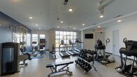 Located at 1817 Sir Tyler Dr in Wilmington, NC, this fitness center offers a well-equipped, single-level workout facility designed to support a full range of exercise routines. The space is bathed in natural light through large floor-to-ceiling windows that look out onto the surrounding exterior, creating an open and energizing atmosphere. Recessed lighting throughout the ceiling ensures consistent illumination during any time of day, while multiple ceiling fans promote comfortable airflow during workouts. The cardio area is outfitted with a lineup of treadmills and elliptical machines, each equipped with individual display screens for tracking performance. A dedicated free weight section features a comprehensive dumbbell rack with a wide range of weights, accompanied by an adjustable workout bench for strength training versatility. Additional strength and conditioning equipment, including cable machines and specialty resistance units, round out the offerings. A wall-mounted flat-screen television adds an entertainment element to the workout environment. The clean, light-colored walls and smooth flooring contribute to a modern, professional aesthetic. The space is organized efficiently, allowing users to move freely between equipment zones while maintaining a spacious feel throughout.