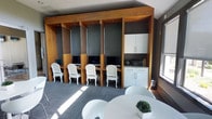 This single-floor space combines functional design with a polished, contemporary aesthetic. The most prominent feature is a custom-built row of individual workstations framed in warm-toned wood cabinetry, each equipped with a computer monitor and a elegantly styled white chair with carved legs, blending classic detailing with modern utility. The built-in shelving units divide each station, offering a degree of privacy while maintaining an open feel throughout the room.

To the side, white upper and lower cabinetry with clean-lined hardware provides additional storage, complemented by a granite-topped counter housing office equipment such as a printer. The walls are finished in a deep charcoal gray, creating a strong contrast against the natural wood tones and bright white furnishings. Patterned gray carpet tiles cover the floor, adding texture underfoot.

A round white table with matching white chairs occupies the foreground, offering a collaborative or casual seating area. Natural light filters in through windows along the perimeter, casting sunlight across the space and enhancing the overall bright, productive ambiance.