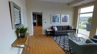 This single-floor residential space offers a thoughtfully designed layout that balances comfort and functionality. The living area serves as the heart of the unit, featuring warm-toned hardwood flooring that runs throughout, creating a cohesive and inviting atmosphere. Neutral, light beige walls provide a clean backdrop that enhances the sense of openness and airiness in the space.

The living room is appointed with a large, tufted dark grey sectional sofa positioned atop a bold geometric-patterned area rug, anchoring the seating area with style. A sleek glass-top side table and accent greenery add refined touches to the arrangement. Two framed art pieces with vibrant color accents are displayed prominently on the main wall, contributing a modern aesthetic.

The space flows naturally through a wide doorway into the kitchen and hallway beyond, where dark cabinetry and contemporary finishes are visible. This open-concept connection between the living and kitchen areas promotes ease of movement and a sense of expansiveness throughout. Ceiling vents suggest a well-integrated climate control system, and the overall layout reflects efficient, modern residential design. The result is a clean, contemporary living environment that is both practical and visually appealing.