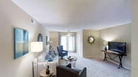 This single-floor residence offers a thoughtfully designed living space that combines comfort and functionality. The living room serves as the focal point of the home, showcasing wall-to-wall plush carpeting that adds warmth underfoot and a soft, neutral color palette across the walls that creates a calm and inviting atmosphere throughout.

Natural light flows generously into the living area through a set of glass patio doors dressed with patterned curtains, offering a bright and airy feel. A ceiling fan positioned centrally above the seating area provides both comfort and a subtle architectural detail. The room accommodates a well-appointed seating arrangement, including a sofa, an accent armchair, and a leather chair, all centered around a round side table — making it an ideal gathering space.

Decorative wall accents, including a starburst mirror and framed artwork, add visual interest to the neutral walls, while floor lamps contribute additional ambient lighting. The textured ceiling adds a classic residential character to the space. The open layout allows for easy flow between areas, enhancing the overall sense of spaciousness. This home is a comfortable, move-in ready space well-suited for everyday living.