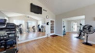 This single-floor fitness center offers a thoughtfully designed workout environment spread across interconnected exercise zones. The space features warm hardwood flooring throughout, vaulted ceilings that create an open and airy atmosphere, and neutral-toned walls that contribute to a clean, motivating aesthetic.

The main exercise area is anchored by a large wall-to-wall mirror that visually expands the space and allows for form-checking during workouts. A wall-mounted flat-screen television provides entertainment during exercise sessions. The room is equipped with a diverse array of cardio machines, including treadmills, an elliptical trainer, a stationary bike, and a stair climber, as well as a multi-station cable strength training system and free weights.

Natural light pours in through multiple large windows fitted with vertical blinds, offering views of the surrounding outdoor environment. A ceiling fan ensures comfortable air circulation, and a standing floor fan provides additional airflow throughout the space. A glass-paned interior door with a classic French-style grid design connects the workout areas, adding a touch of architectural character.

The layout flows naturally between zones, allowing multiple users to exercise simultaneously without congestion. An exterior door with access to the outdoors enhances ventilation and convenience, making this fitness center a comprehensive and inviting space for a full range of workout routines.