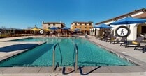 This single-story community amenity space in Brentwood, California (94513) is thoughtfully designed to provide residents with a welcoming outdoor retreat. The centerpiece of the property is a well-maintained swimming pool, enclosed by a sturdy black wrought iron security fence equipped with a gated entry. The pool area features a generous concrete deck, a covered pergola structure, and multiple yellow shade umbrellas, offering ample space for lounging and recreation under the California sun.

Adjacent to the pool enclosure is a covered outdoor patio area finished with smooth concrete flooring and crisp white stucco walls. This shaded space is furnished with wicker lounge seating, including a chaise lounge and a multi-seat sofa adorned with colorful accent pillows, creating a comfortable setting for relaxation. An overhead infrared heater is mounted to the ceiling, extending the usability of this outdoor space into cooler seasons.

Large sliding glass doors with dark frames and etched branding connect the covered patio to an interior space, allowing natural light to flow freely between the two areas. Views of the pool, surrounding landscaping, and open blue skies are visible throughout, reinforcing the bright, open-air ambiance that defines this amenity space.