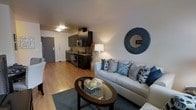 This single-floor unit showcases a thoughtfully designed open-concept layout where the living area and kitchen flow seamlessly into one another. The kitchen is outfitted with dark espresso cabinetry that extends to the upper walls, providing ample storage space. Stainless steel appliances, including a microwave and range, complement the dark stone countertops, while a double sink adds functional convenience. The cabinetry's rich tones create a striking contrast against the light-colored walls and warm hardwood-style flooring that runs throughout the space.

The living area is generously proportioned, centered around a light grey sofa dressed with an array of decorative pillows in navy, teal, and grey tones. A round glass-top coffee table with a dark wood base anchors the seating arrangement over a textured area rug. A decorative round mirror with a metallic frame serves as a focal point on the main wall, adding depth and visual interest. A floor lamp with a classic white shade provides warm ambient lighting, complementing the overhead flush-mount fixtures. The entry door is visible from the living space, reinforcing the practical, connected nature of the layout.