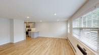 Located at Norwich Gate in East Norwich, NY, this single-floor residence presents a clean and contemporary design with a thoughtfully open layout. The main living area is expansive and airy, featuring light-toned hardwood flooring, recessed lighting, and crisp white walls that create a bright, welcoming ambiance. Large windows fitted with blinds allow generous natural light to fill the space while offering a view of the surrounding greenery.

The kitchen is seamlessly integrated into the open living area via a peninsula counter with a white quartz surface. Gray shaker-style cabinetry lines the walls, complemented by a full suite of stainless steel appliances, including a refrigerator, electric range, and over-the-range microwave. The hardwood flooring continues throughout, maintaining a cohesive feel.

A carpeted bedroom offers a quieter, softer environment, complete with recessed lighting, a window with natural light, and wall-mounted climate control. Additional rooms visible in the layout provide flexible living or storage options, with double closet doors adding practical storage.

The bathroom is finished with classic white subway tile surrounding a soaking tub and shower combination, accented by brushed nickel fixtures for a polished, timeless look. Throughout the residence, the design emphasizes simplicity, functionality, and modern comfort.