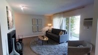 This single-story space showcases a well-appointed living room designed with both comfort and style in mind. The room is anchored by a dark charcoal sofa accented with decorative throw pillows, positioned atop a plush, round area rug that adds texture and warmth to the wood-look flooring beneath. A sleek oval coffee table serves as a functional centerpiece, complemented by a coordinating accent chair.

Natural light flows generously into the space through a large sliding glass door dressed with sheer white curtains, offering a view of lush greenery outdoors and creating an airy, open atmosphere. A floor lamp in the corner provides additional ambient lighting for the evenings.

The walls are kept neutral and clean, providing a versatile backdrop for a set of four framed geometric art pieces arranged in a grid formation, adding a contemporary touch to the décor. The textured ceiling adds subtle architectural character to the room.

Overall, the space presents a cohesive, modern aesthetic with a comfortable, inviting ambiance, ideal for relaxed everyday living.