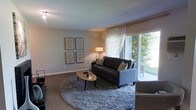 This single-story space showcases a well-appointed living room designed with both comfort and style in mind. The room is anchored by a dark charcoal sofa accented with decorative throw pillows, positioned atop a plush, round area rug that adds texture and warmth to the wood-look flooring beneath. A sleek oval coffee table serves as a functional centerpiece, complemented by a coordinating accent chair.

Natural light flows generously into the space through a large sliding glass door dressed with sheer white curtains, offering a view of lush greenery outdoors and creating an airy, open atmosphere. A floor lamp in the corner provides additional ambient lighting for the evenings.

The walls are kept neutral and clean, providing a versatile backdrop for a set of four framed geometric art pieces arranged in a grid formation, adding a contemporary touch to the décor. The textured ceiling adds subtle architectural character to the room.

Overall, the space presents a cohesive, modern aesthetic with a comfortable, inviting ambiance, ideal for relaxed everyday living.