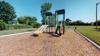 Located at 2209 Wabash Ct W in Columbus, Ohio, this outdoor community playground offers a welcoming recreational space designed for active play. The centerpiece of the area is a multi-level play structure finished in a bold dark metal frame, featuring covered platforms that provide shade, an open slide, climbing elements, and connecting staircases. The structure is anchored on a generous wood chip surface, which serves as both a safety cushion and a defined play zone. Surrounding the playground is a broad expanse of open, well-kept green lawn, providing ample room for free-range outdoor activities. Mature trees line the perimeter of the space, offering natural shade and a pleasant, park-like atmosphere. A waste receptacle is conveniently positioned near the play area, reflecting thoughtful maintenance of the grounds. The open sky and lush greenery create a bright, inviting environment suited for children and families. The overall design emphasizes accessibility and safety, with clearly delineated play and open grass areas working together to create a functional and enjoyable community outdoor space.