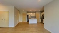 This single-floor space features an open-concept layout that seamlessly integrates the living area and kitchen into one cohesive environment. Light hardwood flooring spans the entirety of the space, contributing to a bright, clean aesthetic. The kitchen is a central highlight, anchored by a white island with a built-in sink and pendant lighting overhead. White cabinetry lines the kitchen walls, paired with stainless steel appliances including a refrigerator, stove, and microwave. Recessed lighting is installed throughout the ceiling, providing even illumination across the open floor plan. The walls are finished in a neutral gray-beige tone, complementing the modern design. Two interior doors are visible — one leading to what appears to be a utility or storage area and another to an additional room — suggesting a functional layout beyond the main living space. The open arrangement between the kitchen and living area creates a generous, versatile space well-suited for both everyday living and entertaining. Overall, the design emphasizes clean lines, practicality, and a contemporary aesthetic with a light, airy ambiance.