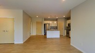 This single-floor space features an open-concept layout that seamlessly integrates the living area and kitchen into one cohesive environment. Light hardwood flooring spans the entirety of the space, contributing to a bright, clean aesthetic. The kitchen is a central highlight, anchored by a white island with a built-in sink and pendant lighting overhead. White cabinetry lines the kitchen walls, paired with stainless steel appliances including a refrigerator, stove, and microwave. Recessed lighting is installed throughout the ceiling, providing even illumination across the open floor plan. The walls are finished in a neutral gray-beige tone, complementing the modern design. Two interior doors are visible — one leading to what appears to be a utility or storage area and another to an additional room — suggesting a functional layout beyond the main living space. The open arrangement between the kitchen and living area creates a generous, versatile space well-suited for both everyday living and entertaining. Overall, the design emphasizes clean lines, practicality, and a contemporary aesthetic with a light, airy ambiance.